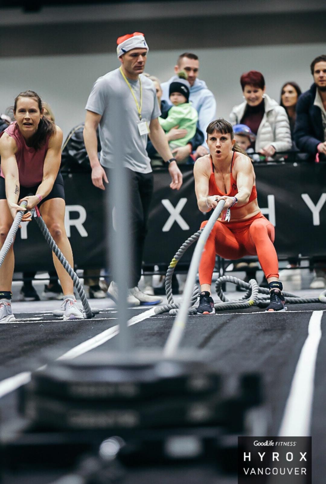 Two women perform battle rope exercises at a Goodlife Fitness Hyrox event in Vancouver with spectators watching.
